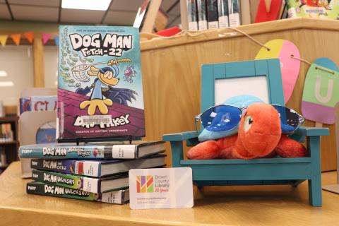 A library card rests next to a stuffed turtle sitting in a toy chair near a stack of Dogman books.