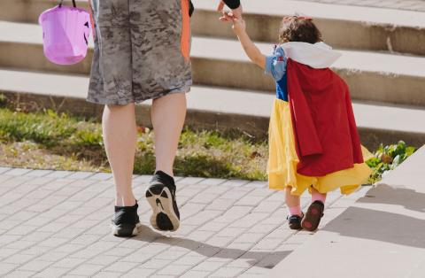 An adult and child are walking holding hands. The child is in a Snow-White costume. The adult is holding a purple bucket pail. They are walking away from the camera.