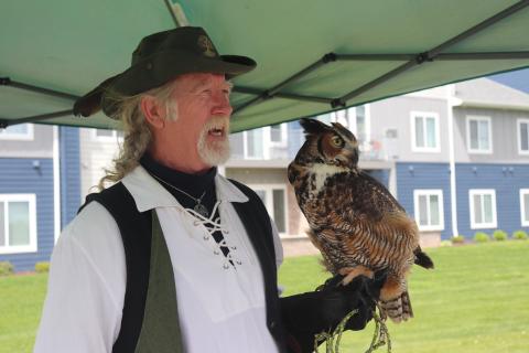 Man with long gray hair and goatee in a green feathered hat and black vest over a white shirt holding a Great Horned Owl on a gloved hand.