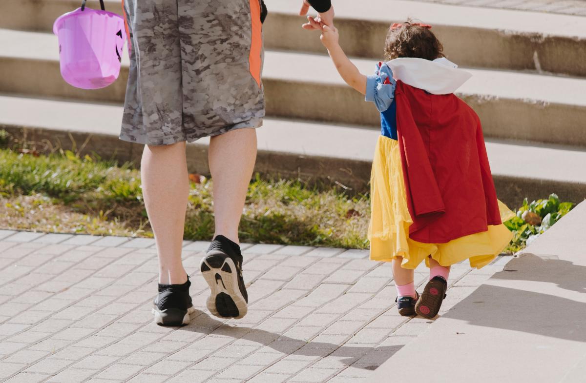 An adult and child are walking holding hands. The child is in a Snow-White costume. The adult is holding a purple bucket pail. They are walking away from the camera.