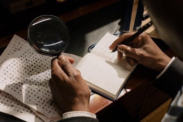 Man's hands holding a magnifying glass over a coded message while writing in a small notebook